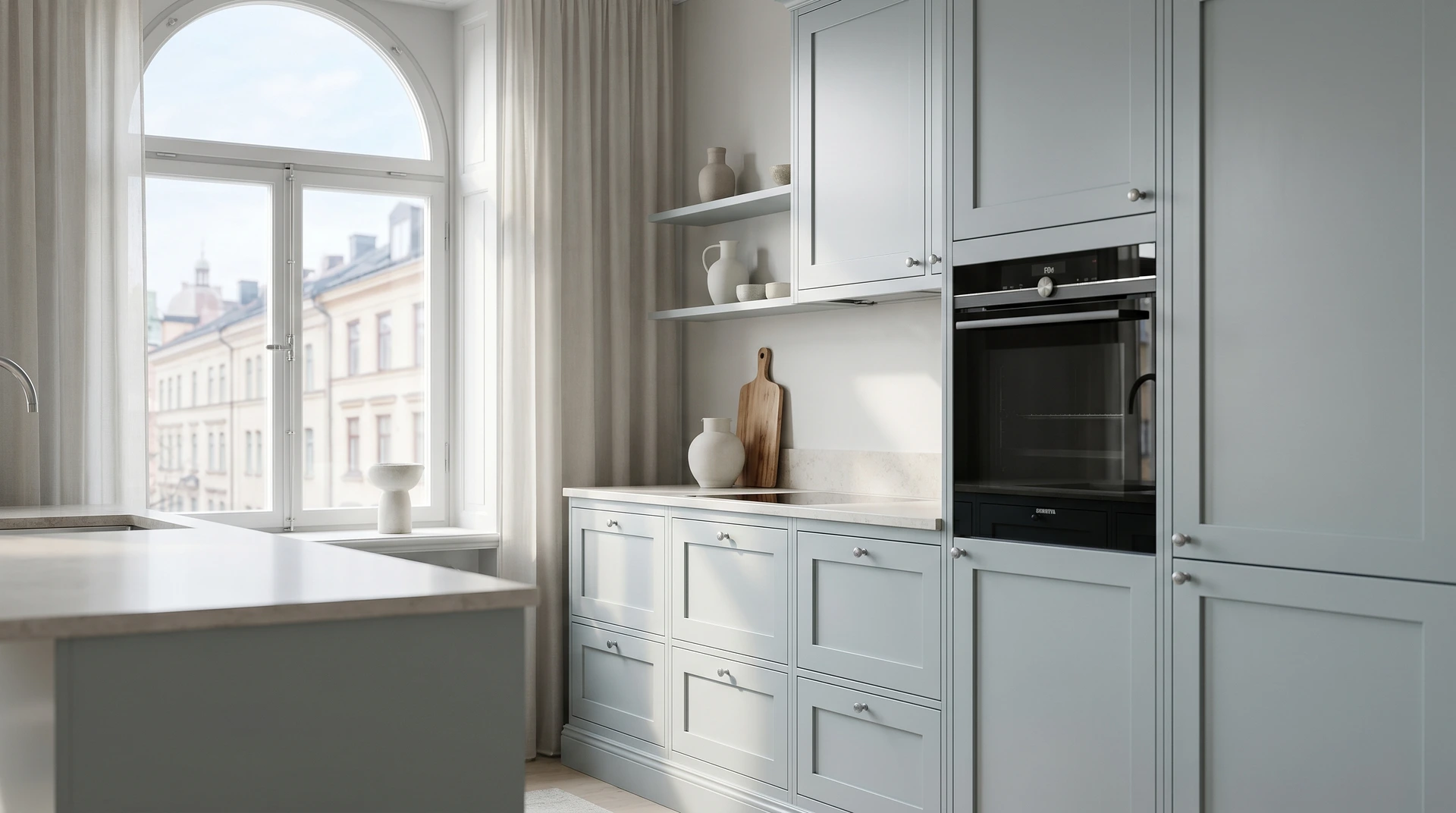 Ljust kök med gråa skåp, inbyggd ugn och stort rundbågefönster.